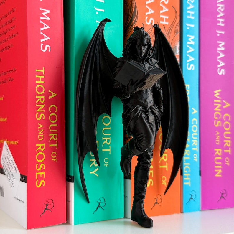 Pu&ograve; includere: Una statuetta nera di una figura alata che legge un libro, posizionata tra dorsi di libri colorati. I libri hanno titoli come "A Court of Thorns and Roses" e "A Court of Wings and Ruin". La statuetta ha le ali spiegate e indossa stivali.
