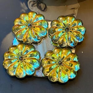May include: Four iridescent flower-shaped decorative objects. Each flower has multiple petals and a dark centre. The flowers are arranged in a square on a reflective surface, with a dark background.