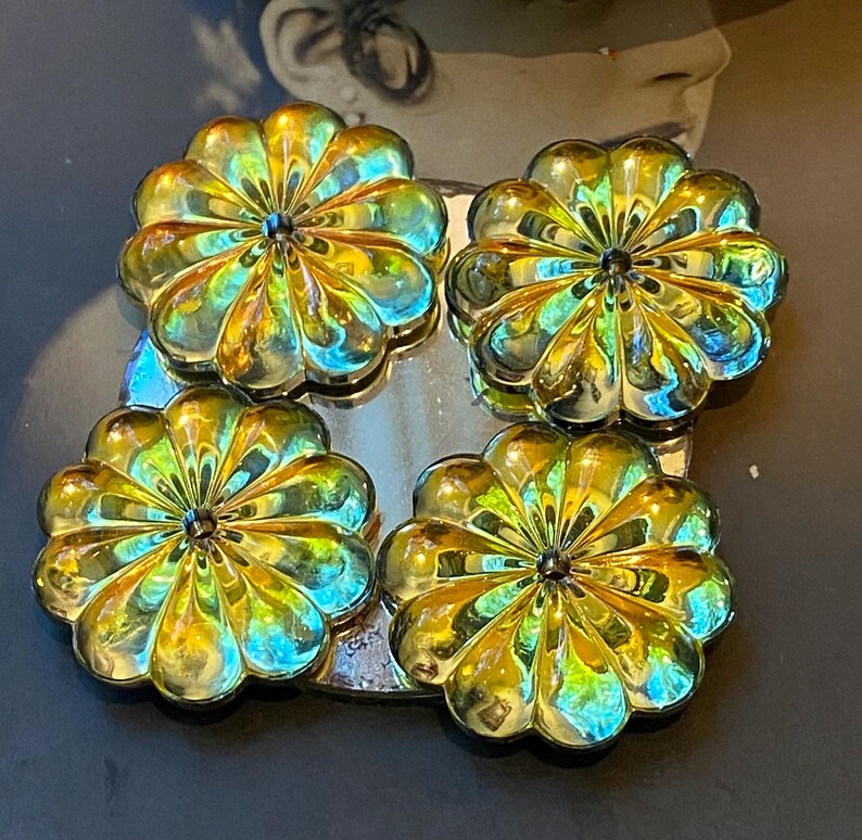 May include: Four iridescent flower-shaped decorative objects. Each flower has multiple petals and a dark centre. The flowers are arranged in a square on a reflective surface, with a dark background.
