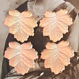 May include: A set of four off-white, leaf-shaped decorative items with a smooth, matte finish and detailed vein patterns. Each piece features a symmetrical, multi-lobed design with scalloped edges, arranged in a two-by-two grid formation.