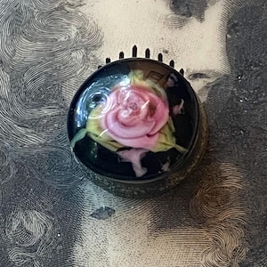 May include: A round, black glass paperweight featuring an embedded pink rose design with light green leaves. The object has a smooth, domed surface and a dark, textured base. The rose is centered within the clear, dark glass sphere.