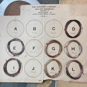 May include: Vintage sample card from The Hakuzen Company, featuring silver and gold thread and tinsel. The card displays circular samples labeled A through L, each with a different color and texture. The text on the card reads "Silver & Gold Thread & Tinsels."