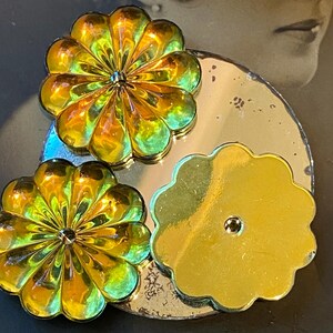May include: Close-up of three flower-shaped decorative objects. Two are iridescent with yellow, orange, and green hues, and the third is a solid gold colour. They are arranged on a silver-coloured, circular base.
