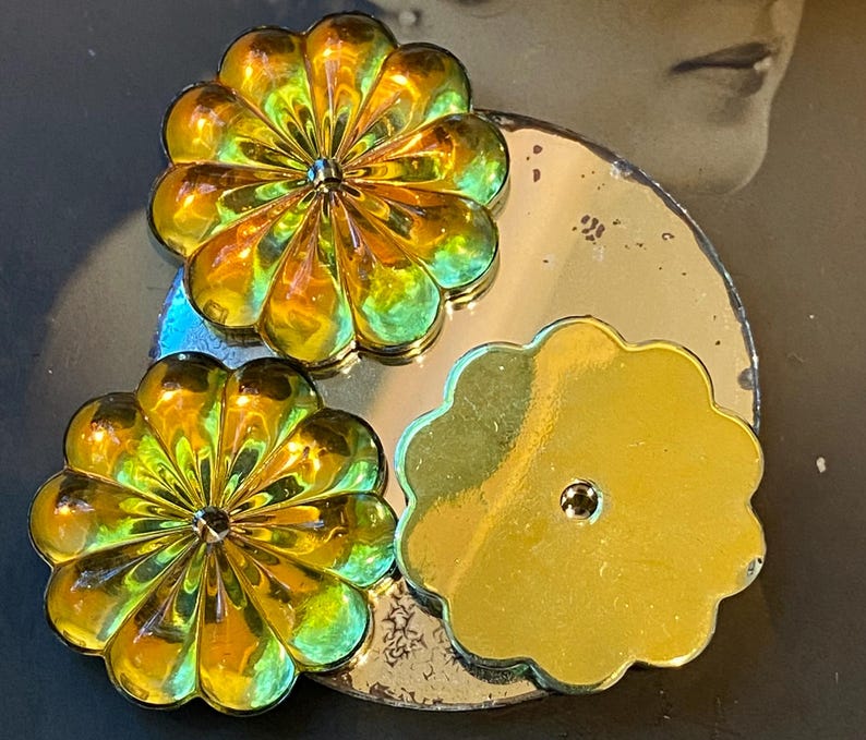 May include: Close-up of three flower-shaped decorative objects. Two are iridescent with yellow, orange, and green hues, and the third is a solid gold colour. They are arranged on a silver-coloured, circular base.