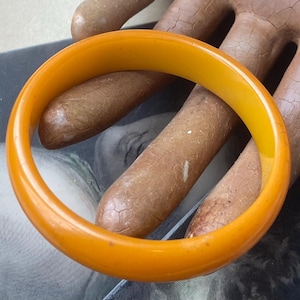 May include: A round, amber-colored bangle bracelet. The bracelet is a solid, smooth band of a warm, golden-orange hue. The jewelry is a simple, classic design, suitable for everyday wear. The bracelet is approximately 0.75 inches wide.
