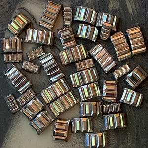 May include: A collection of small, rectangular, silver-toned metal beads with a faceted, stepped design. The beads are scattered across a textured, dark background, reflecting light and creating highlights.