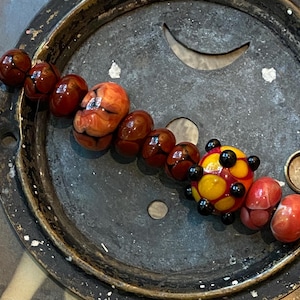 May include: A strand of glass beads featuring a variety of shapes and colors, including deep red, marbled orange, and a prominent yellow sphere with black dots. The beads are strung on a thin metal wire, showcasing a mix of smooth and textured surfaces.