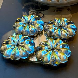 May include: Four iridescent, flower-shaped decorative objects. Each object has a central dark spot and petal-like edges, reflecting blue, green, and gold hues. The objects are arranged on a reflective surface, creating a mirrored effect.