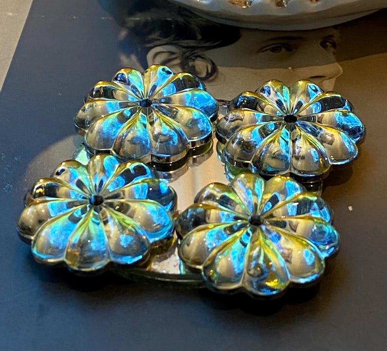 May include: Four iridescent, flower-shaped decorative objects. Each object has a central dark spot and petal-like edges, reflecting blue, green, and gold hues. The objects are arranged on a reflective surface, creating a mirrored effect.
