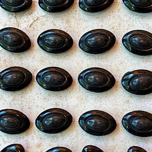 May include: A collection of 20 black oval-shaped buttons with a raised center. The buttons are arranged in rows on a white surface.