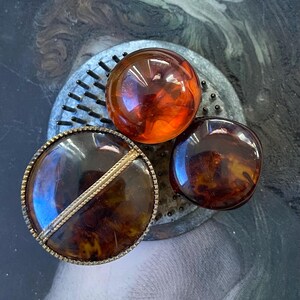 以下が含まれることがあります： 茶色と琥珀色のプラスチックでできたヴィンテージボタン3つ。ボタンは丸く、滑らかで光沢のある仕上がりです。1つのボタンは、縁に金色のトリムが付いています。