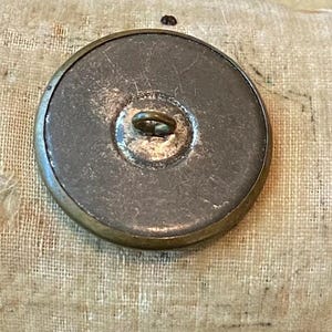 May include: A large, antique button with a tarnished brass rim and a dark, textured center. The button has a metal loop on the back for sewing. The button is round and appears to be vintage.