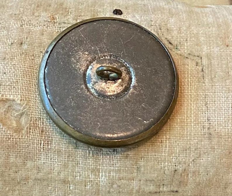 May include: A large, antique button with a tarnished brass rim and a dark, textured center. The button has a metal loop on the back for sewing. The button is round and appears to be vintage.