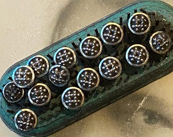 May include: A rectangular, teal-colored metal brush base with rounded ends, featuring numerous upright metal bristles. Attached to the bristles are several circular, dark-toned metal caps, each decorated with a cluster of small, raised silver studs.