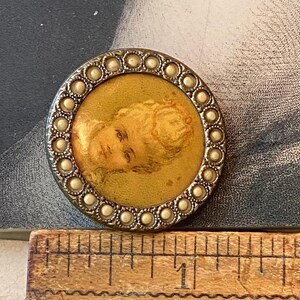 May include: A round, antique button with a portrait of a woman. The button has a silver-colored metal frame with a border of small, pearl-like beads. The portrait is in a gold-colored circle.