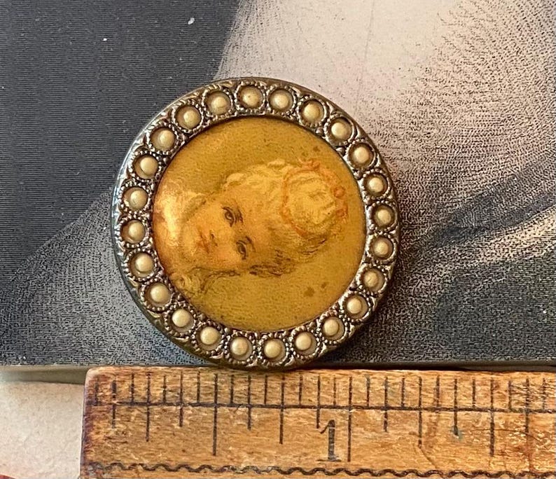May include: A round, antique button with a portrait of a woman. The button has a silver-colored metal frame with a border of small, pearl-like beads. The portrait is in a gold-colored circle.