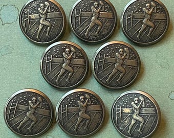 May include: A collection of nine antique silver-toned metal buttons. Each button features a detailed relief of a football player in action, set against a backdrop of a stadium fence. The buttons are round and have a slightly aged appearance.