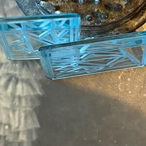 May include: Two rectangular, light blue glass earrings with an abstract geometric design. The earrings have a clear, transparent appearance and are set against a neutral background.