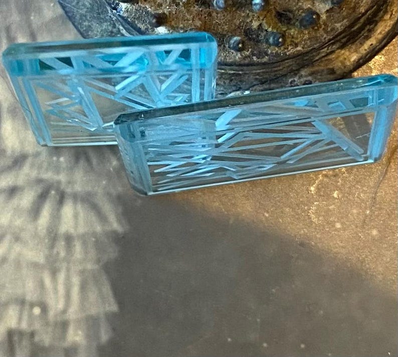 May include: Two rectangular, light blue glass earrings with an abstract geometric design. The earrings have a clear, transparent appearance and are set against a neutral background.
