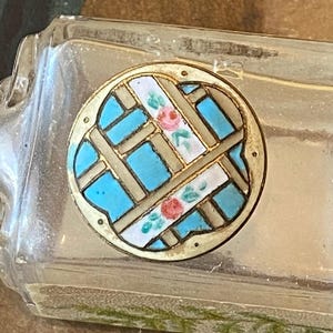 May include: A clear glass perfume bottle with a decorative circular emblem. The emblem has a gold-coloured frame with a blue, white, and tan geometric design, accented with small pink floral details. The bottle has a rectangular shape.