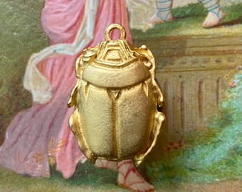 May include: A gold-toned metal scarab beetle pendant featuring a textured, matte finish and a small circular loop at the top for attachment. The beetle design displays detailed wing covers and legs, presented as a singular jewelry component.