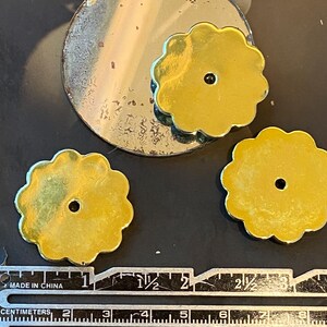 May include: Close-up of three gold-coloured, flower-shaped metal components with a central hole. A circular, tarnished metal piece is also visible. A ruler with centimetre markings is at the bottom. The components appear to be approximately 2.5 cm in diameter.