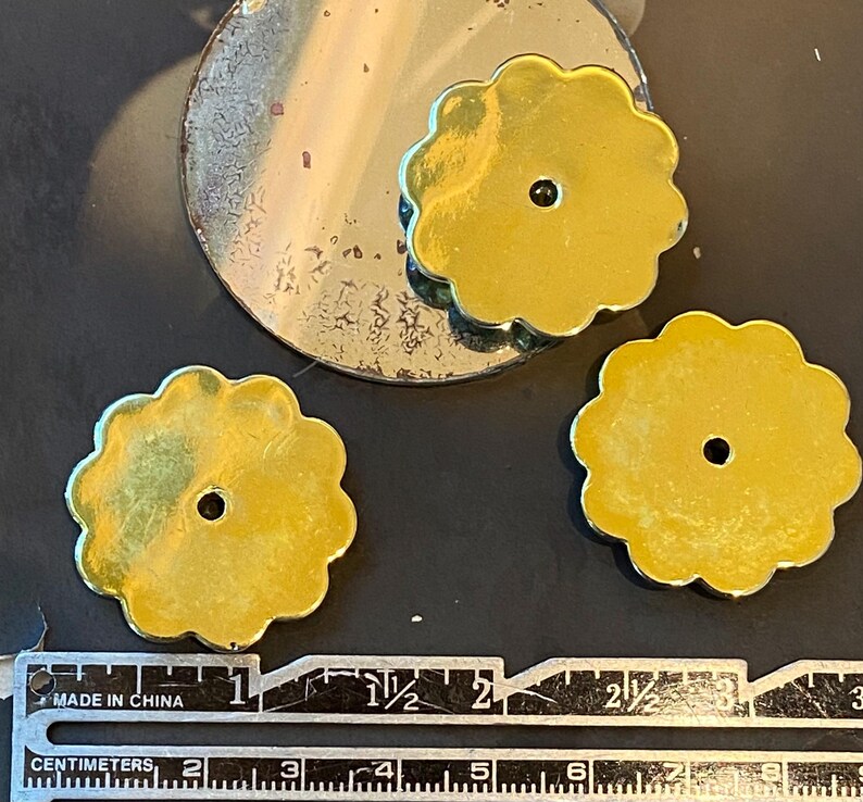 May include: Close-up of three gold-coloured, flower-shaped metal components with a central hole. A circular, tarnished metal piece is also visible. A ruler with centimetre markings is at the bottom. The components appear to be approximately 2.5 cm in diameter.