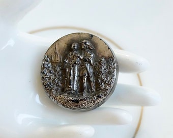 May include: A silver-toned button with a raised design of two figures standing in a forest setting. The button has a smooth, rounded surface.