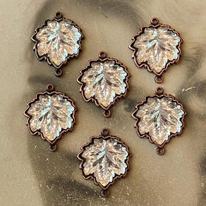 May include: Six leaf-shaped charms featuring clear, faceted glass centers set within copper-toned metal frames. Each charm has a small circular loop at the top and bottom for attachment, showcasing a detailed, textured botanical design.