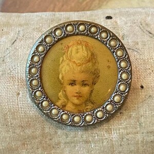 May include: A round, antique button with a portrait of a woman in a yellowed oval. The portrait is surrounded by a silver-toned metal frame with a border of small, white, pearl-like beads. The woman has blonde hair and is wearing a crown.