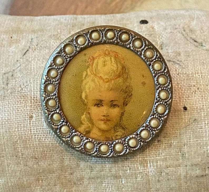 May include: A round, antique button with a portrait of a woman in a yellowed oval. The portrait is surrounded by a silver-toned metal frame with a border of small, white, pearl-like beads. The woman has blonde hair and is wearing a crown.