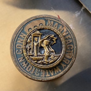 May include: A round, wooden button with a gold-colored design. The design features a small, winged figure standing in front of a building with a clock tower. The text "WALTHAM WATCH COMPANY MANUFACTURED" is written around the edge of the button.