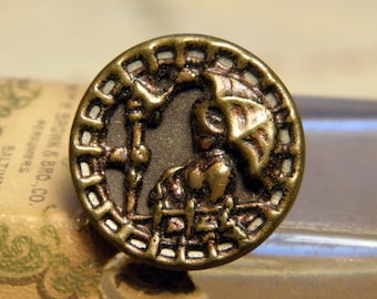 May include: A gold-toned metal button with a raised design of a knight in armor holding a spear. The button has a decorative border with a repeating pattern.