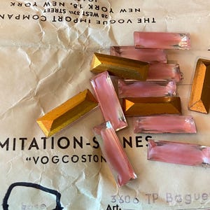 以下が含まれることがあります： ピンクとゴールドの色合いの長方形と台形型のイミテーションストーンのコレクション。ストーンは「The Vogue Import Company」のテキストが入ったヴィンテージペーパーに散らばっています。これらのストーンは、ジュエリー作りやクラフトプロジェクトに適しています。