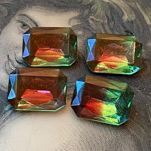 May include: Four iridescent, emerald-cut glass gems. The gems have a color-shifting effect, with hues of green, orange, and blue. The gems are arranged on a patterned surface, showcasing their multifaceted design.