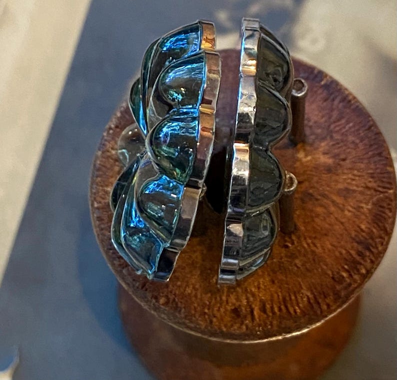 May include: A pair of silver earrings with a distinctive, layered design. The earrings have a dark, iridescent finish reflecting blue and green hues. The earrings are displayed on a wooden surface.