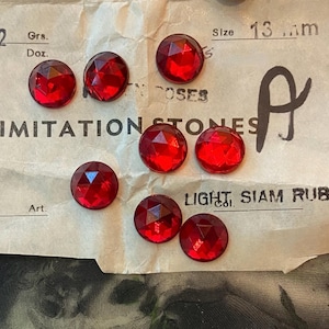 May include: Eight round, faceted red imitation stones arranged on a vintage paper card labeled 13 mm, IMITATION STONES, and LIGHT SIAM RUBY. The stones have a brilliant, multi-faceted surface that reflects light, creating a deep crimson appearance.