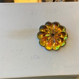 May include: A gold-toned flower brooch with a dark central detail, set against a white surface. The brooch has a layered petal design. The background features faint text and sketches, with the words "Made in Austria" visible.