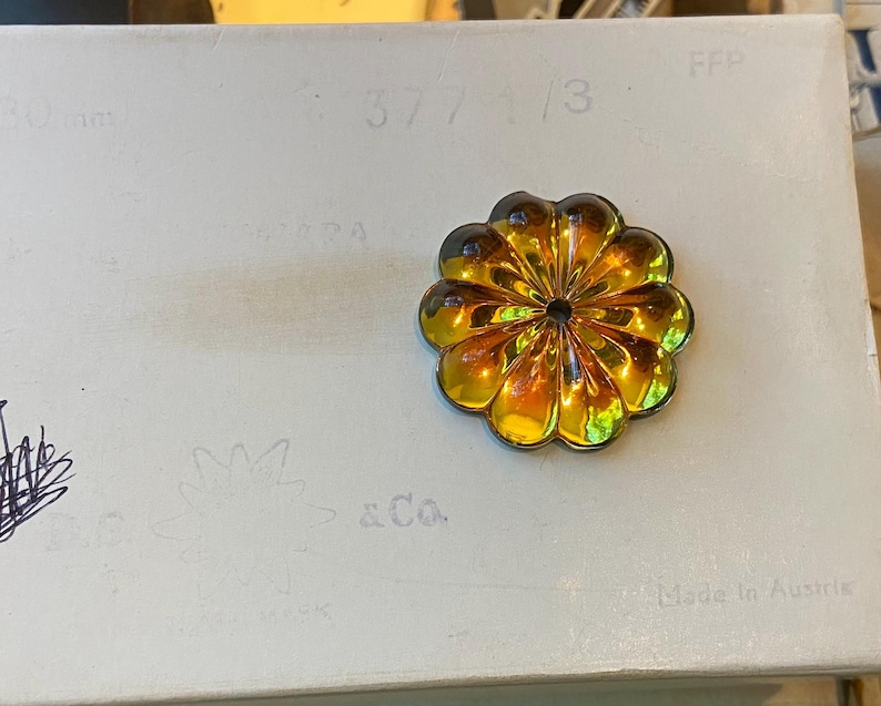 May include: A gold-toned flower brooch with a dark central detail, set against a white surface. The brooch has a layered petal design. The background features faint text and sketches, with the words "Made in Austria" visible.