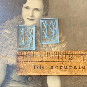 May include: Two light blue rectangular earrings with an abstract geometric design. A wooden ruler with the text "This accurate" is below the earrings. The background is a vintage photograph.