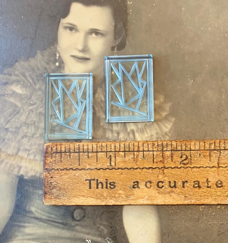 May include: Two light blue rectangular earrings with an abstract geometric design. A wooden ruler with the text "This accurate" is below the earrings. The background is a vintage photograph.