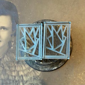 May include: Two rectangular, light blue glass earrings with an Art Deco design. Each earring features a geometric pattern within a rectangular frame. The earrings are displayed on a dark, textured surface, with a vintage photograph in the background.