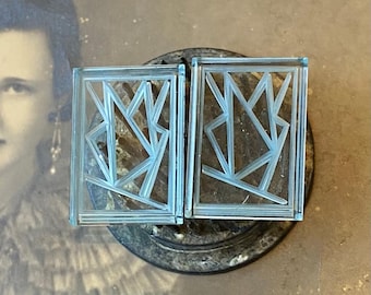 May include: Two rectangular, light blue glass earrings with an Art Deco design. Each earring features a geometric pattern within a rectangular frame. The earrings are displayed on a dark, textured surface, with a vintage photograph in the background.