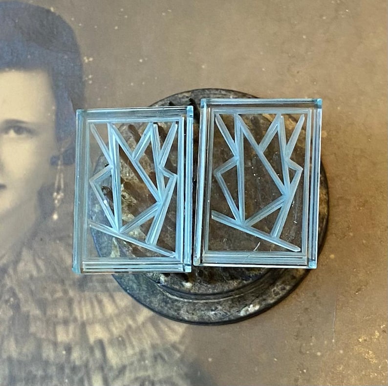 May include: Two rectangular, light blue glass earrings with an Art Deco design. Each earring features a geometric pattern within a rectangular frame. The earrings are displayed on a dark, textured surface, with a vintage photograph in the background.