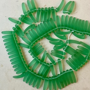 May include: A strand of green glass beads, each bead is shaped like a long, thin, slightly curved leaf. The beads are strung together on a white thread.