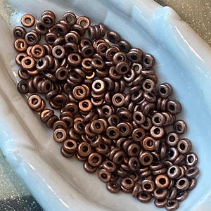 May include: A collection of small, round, metallic copper-colored spacer beads. Each bead features a smooth, circular design with a central hole, exhibiting a consistent dark bronze-toned finish across the entire group of loose jewelry components.