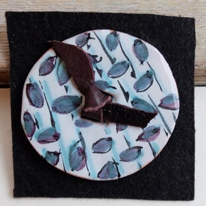 May include: A white ceramic dish with a blue and purple abstract design. A brown leather strap is attached to the dish.