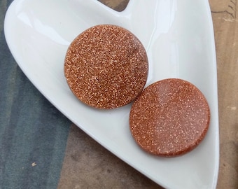 May include: Two round, copper-colored, glittery, sandstone earrings on a white heart-shaped dish.