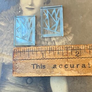 May include: Two small, rectangular, light blue glass pieces with geometric designs. A wooden ruler with the text "This accurate" is below the glass pieces. The glass pieces are approximately 1 inch in length.