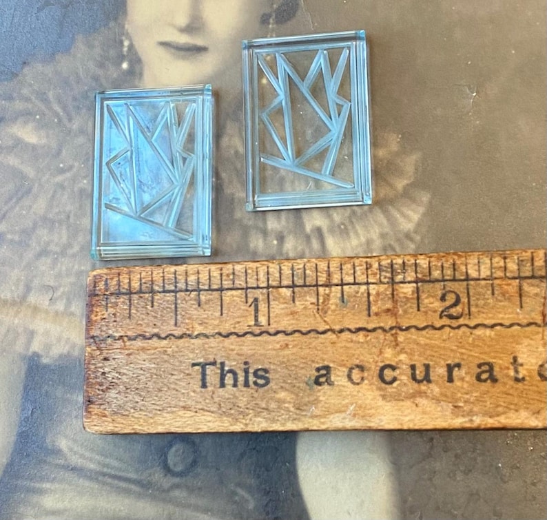 May include: Two small, rectangular, light blue glass pieces with geometric designs. A wooden ruler with the text "This accurate" is below the glass pieces. The glass pieces are approximately 1 inch in length.